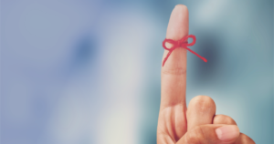 A hand with the index finger pointing up and with a red bow tied around it. This represents and old idea to tie a bow around your finger to remember something.