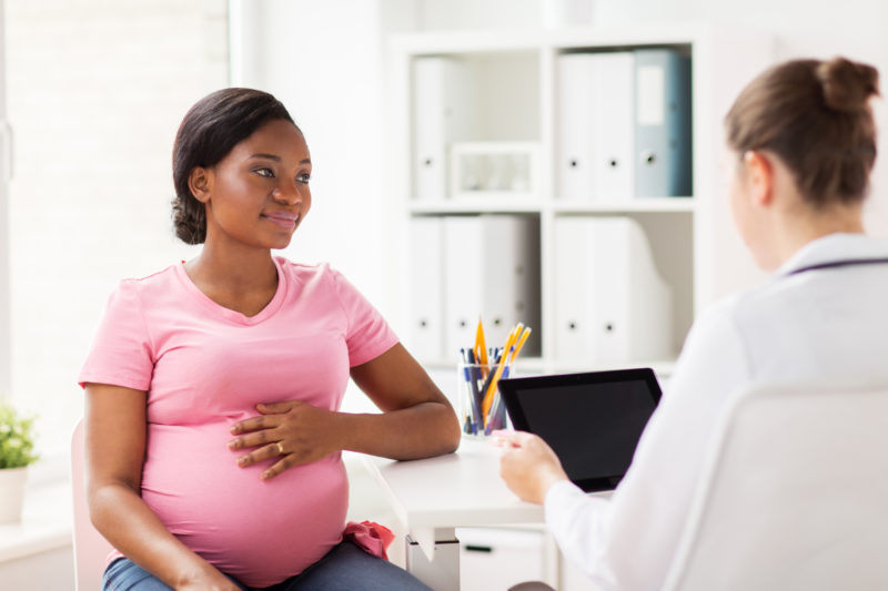 Pregnant woman at the doctors clinic. She is speaking with her doctor. She has a happy expression and has one hand on her belly.