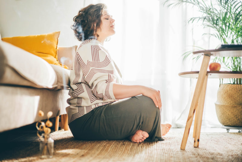 Woman sitting cross-legged on the floor. She has her hands on her knees and her eyes are closed. She is meditating.